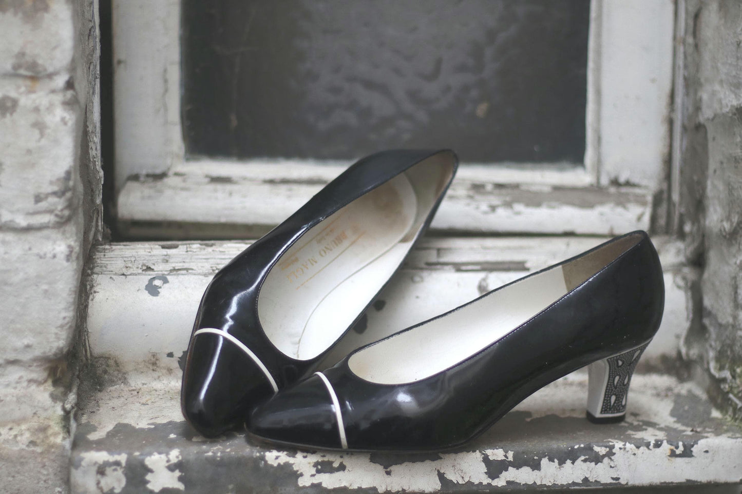 A pair of black Patent Leather Pumps by the brand Bruno Magli photographed outdoors from front view perspective on a white chabby chic window sill . The vintage 80s heels are statement with contrasting white heels and line in front.