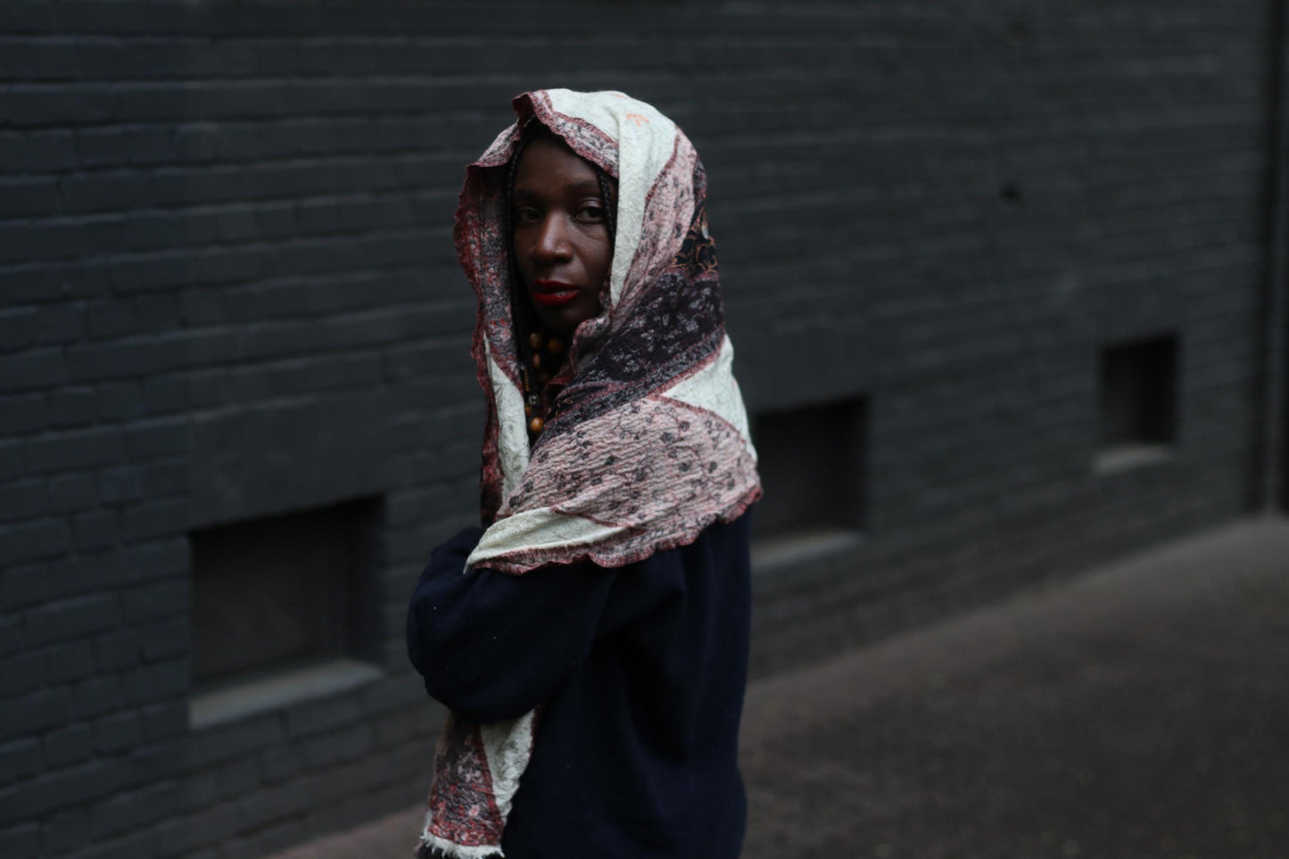 A vintage scarf from the 90s with a distinctive print worn by a female model with beaded braids as a headscarf. The model is standing in front of a dark brick wall.