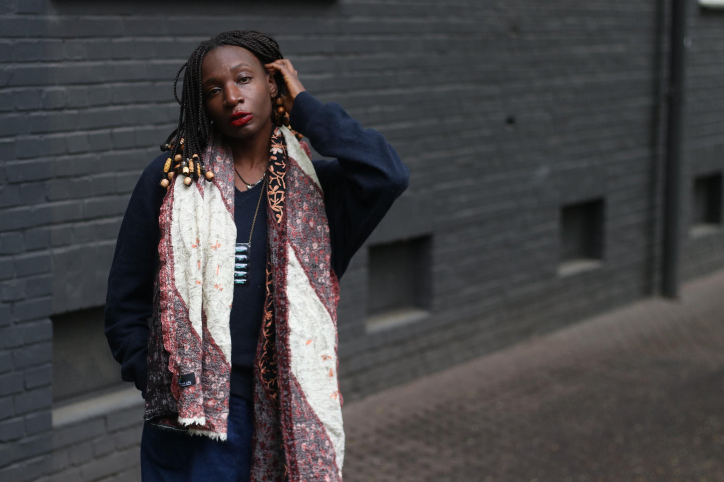 Front view of a retro patterned scarf worn by a female model, the outdoor image, with a textured brick wall in the background, showcases the unique print and color palette of the boho oversized scarf.