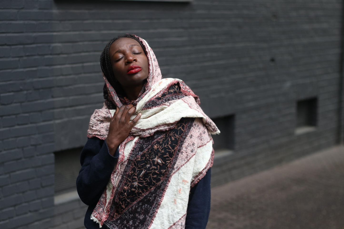 Close-up of a retro patterned scarf worn as a head scarf by a female model, the outdoor image, with a textured brick wall in the background, showcases the unique print and color palette of the boho oversized scarf.