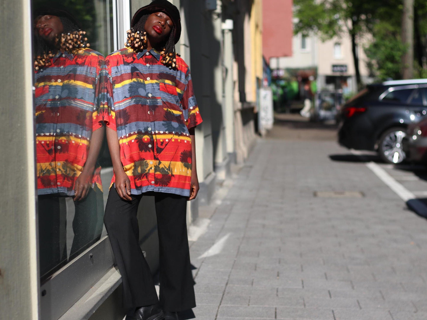 Female model in a vintage Montino tropical floral shirt with bold island print styled with black pants and a slouchy hat photgraphed in an urban setting with buildings and cars in the background.