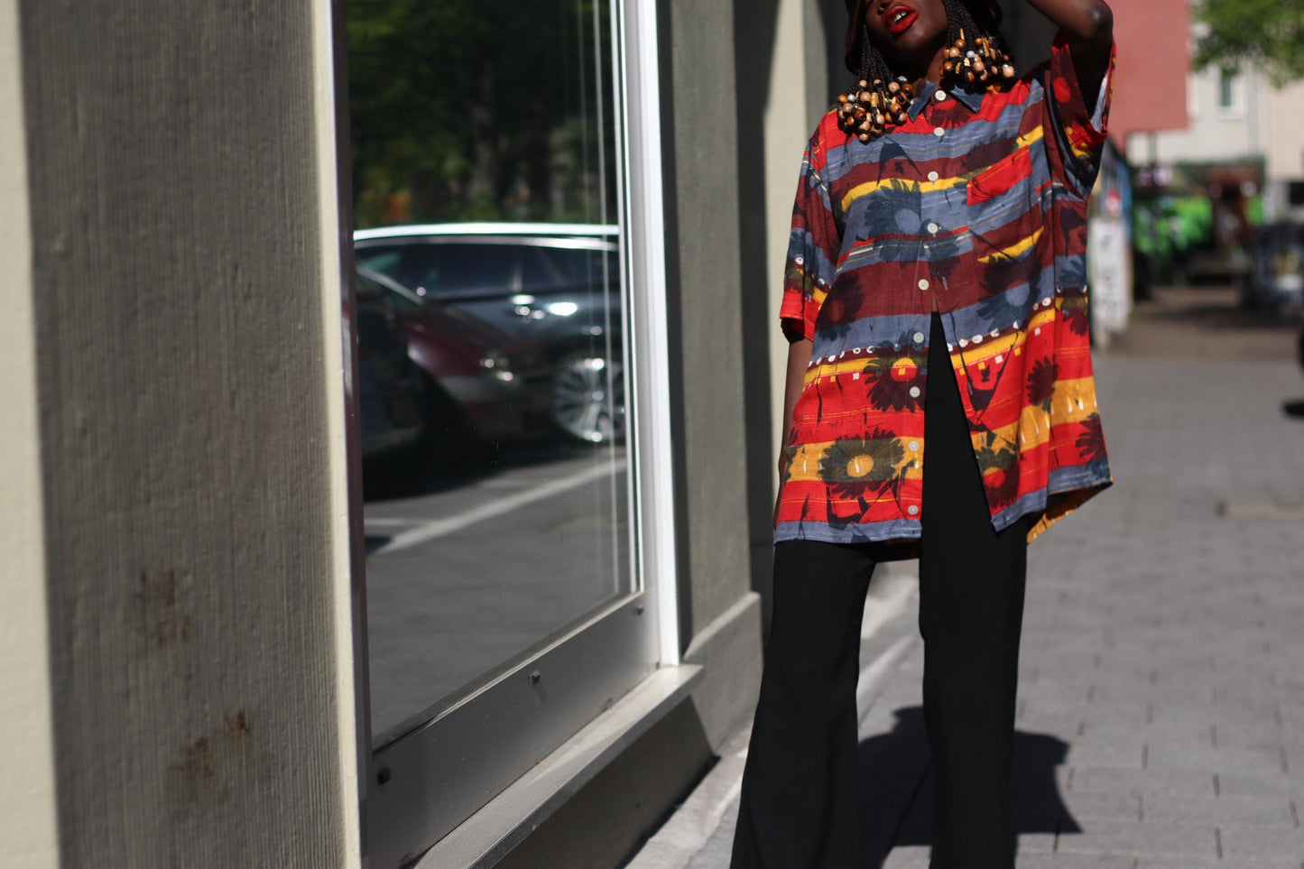 Female model wearing a 90s retro floral print shirt in red and yellow, black trousers, and a hat stands on a paved area next to parked cars. They are positioned between a building with a glass storefront and a row of vehicles.