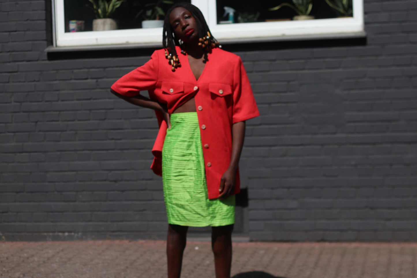 Female model wearing a vintage 90s Short-Sleeve Blazer by Mark Wald with a neon green knee-length skirt photographed in front of a grey brick wall with a white window sill visible. The image showcases the bold V-Neck Jacket as a lightweight choice for suiting up in the summer