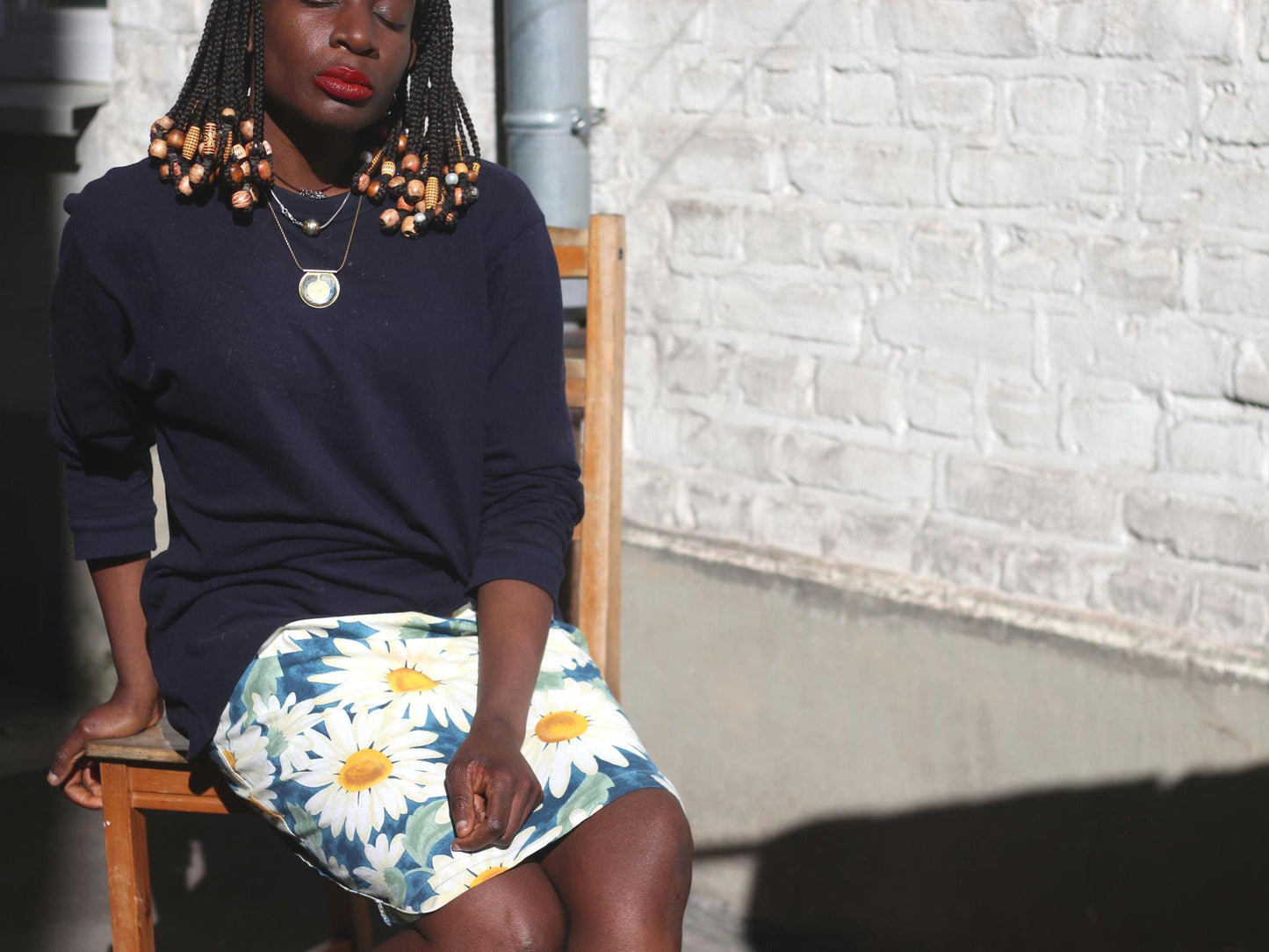 Woman with dark skin and beaded braids wearing a navy top and a vintage 90s daisy-print skirt, seated on a wooden chair against a light-colored wall
