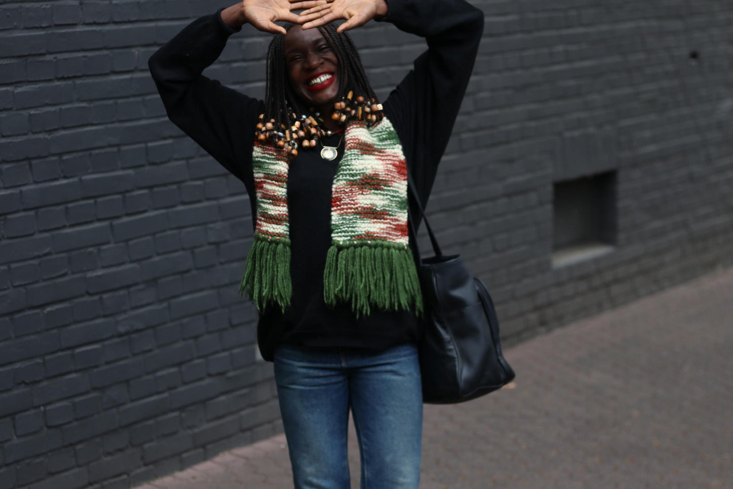 70s handknitted scarf with multicolor yarn and fringe detailing, styled with casual winterwear and big bag on a female model captured outdoors against a grey brick wall
