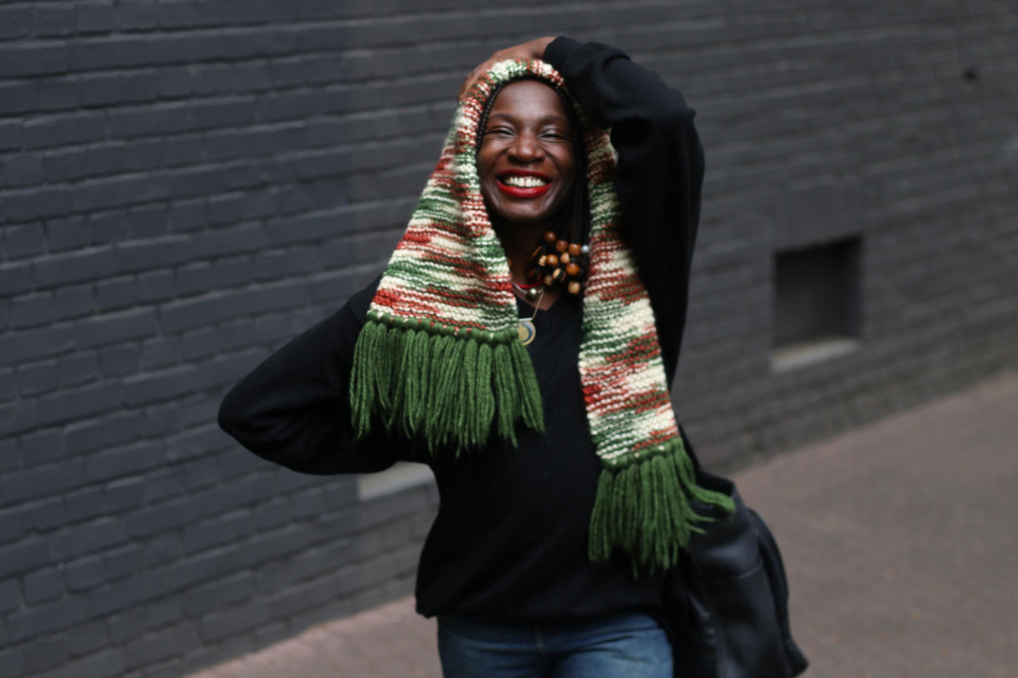 Handmade vintage green knit scarf with chunky tassels worn by a smiling woman ias a head covering . the woman is also wearing a black sweater and big handbag and is standing against a brick wall