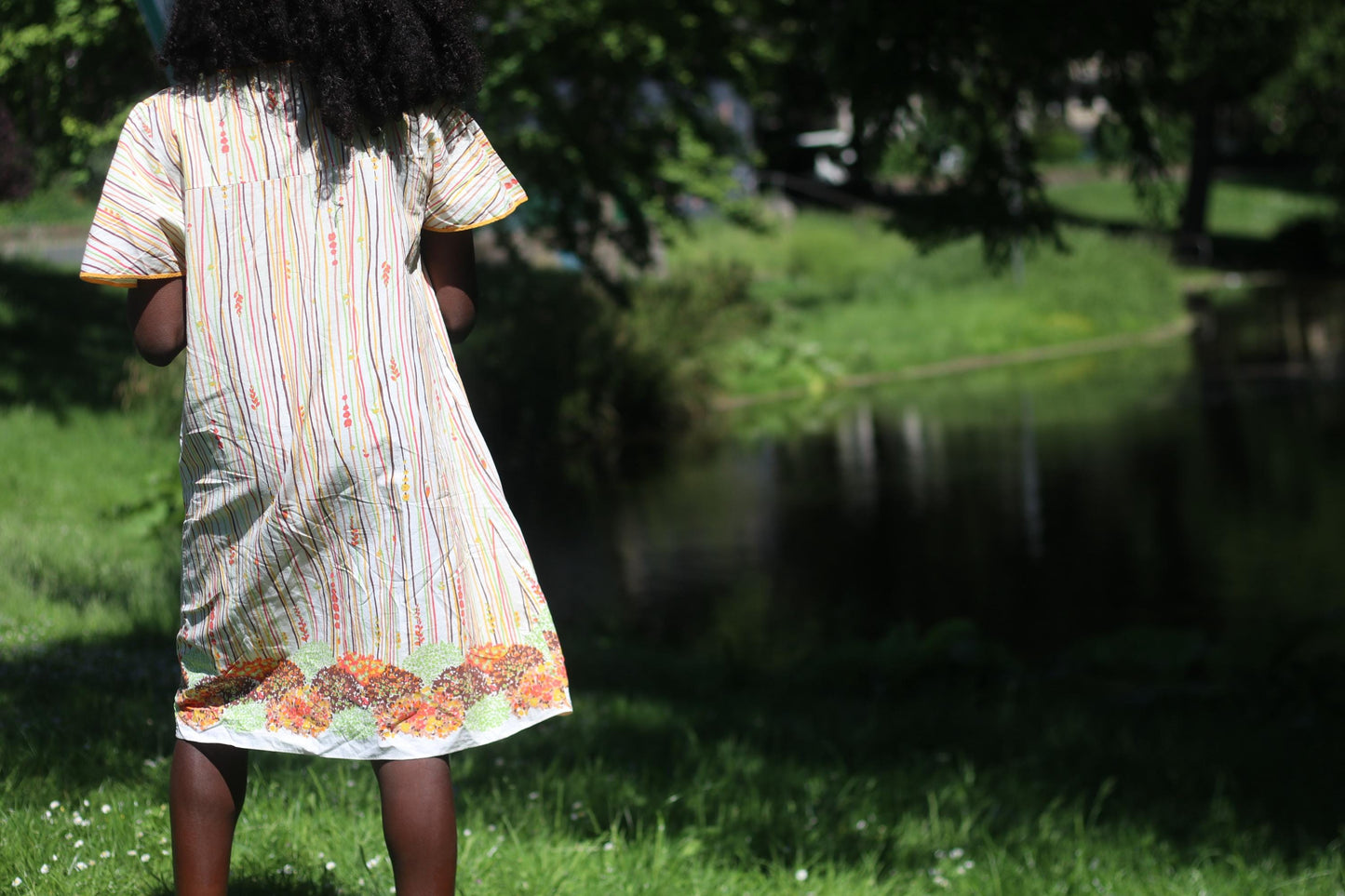 Back view of a vintage boho retro style 70s dress on a female model with curly black hair photographed in front of a lake with plenty of greenery