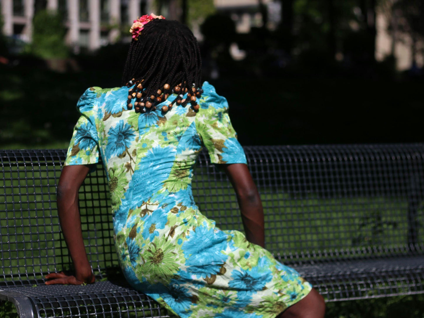 A back view of a female model with dark skin and beaded braids sits on a park bench. They are wearing a vibrant vintage blue and green floral dress with short, puffed sleeves and a colorful floral headpiece.