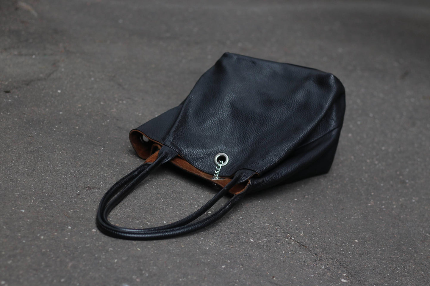 A black, pebbled leather tote bag with a brown lining and silver chain detail, laying on a dark textured surface.