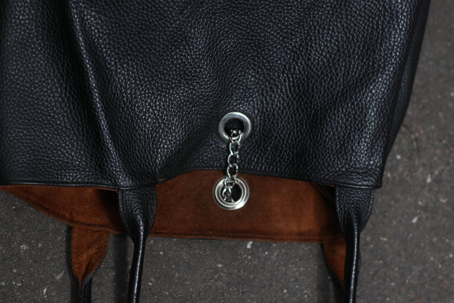 close up of silver chain detail on a vintage black, pebbled leather tote bag with a brown lining laying on a dark textured surface.