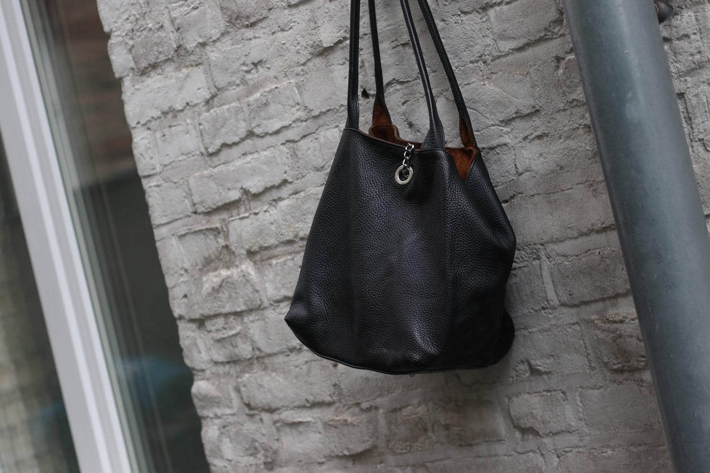 Vintage 90s minimalist legant black pebbled leather shoulder bag with a silver grommet and chain feature hanging against a white bricked wall