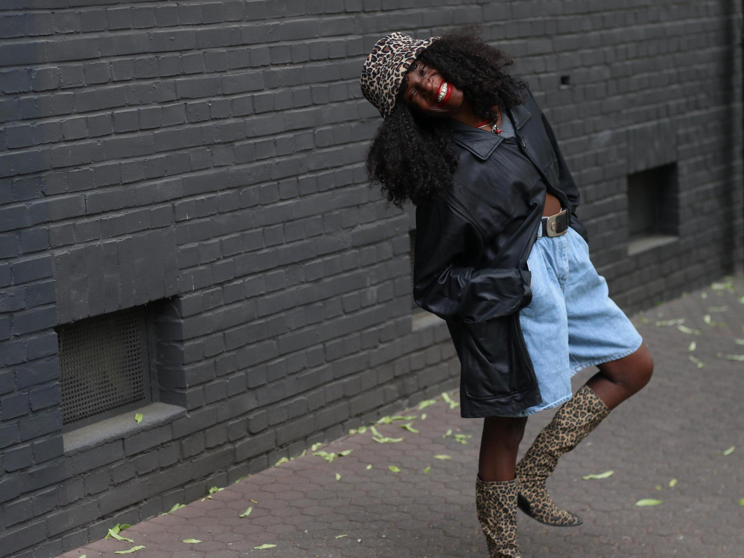 A female model wearing a black vintage oversized 90s leather jacket with a leopard print hat and boots, posing against a dark brick wall.