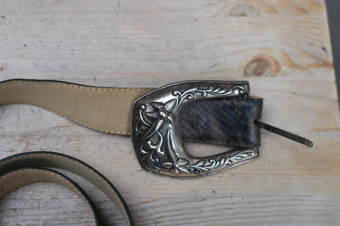Overhead shot of a dark snakeskin-embossed vintage leather western belt with an ornate silver buckle, photographed on a light wooden surface