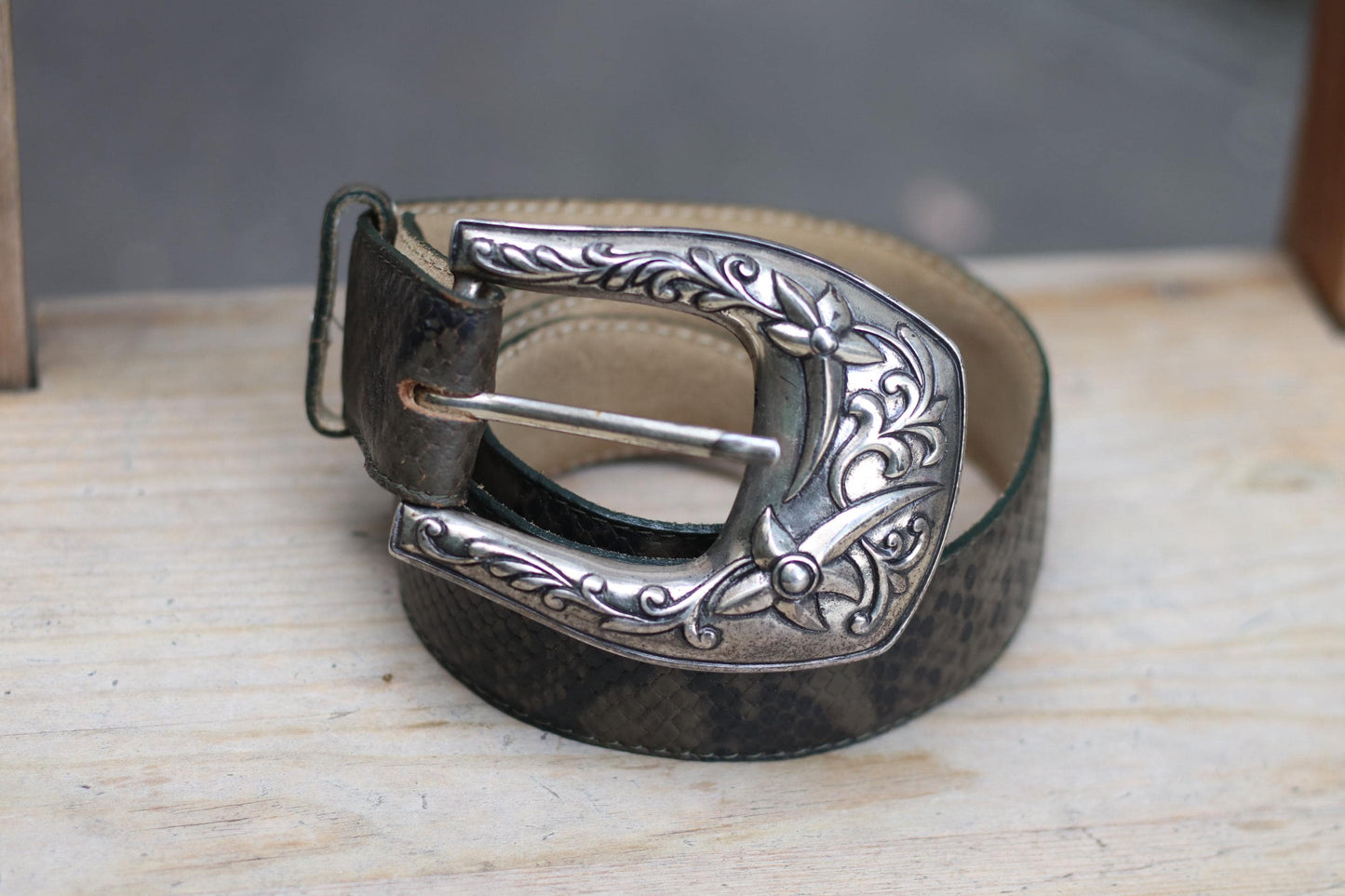 A vintage western belt from the 70s with an ornate silver buckle, coiled on a rustic wooden surface, showcasing the snakeskin-embossed dark leather.