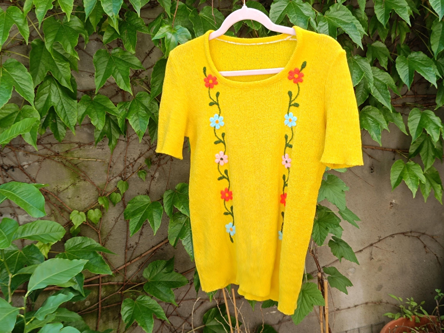 Retro-style vintage yellow short sleeved top with vertical flower embroidery, photographed hanging on a clothing line outdoors against green leafy background