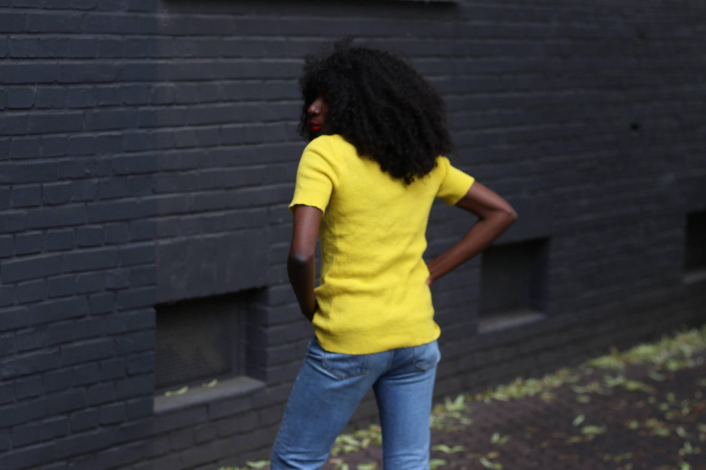 Back view of a female model in a sunny vintage yellow shirt and casual blue jeans photographed outdoors against a grey brick wall.