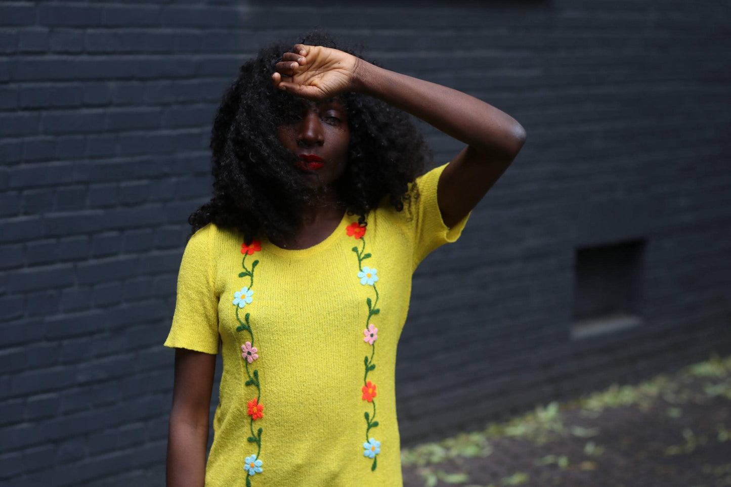 Artistic shot of a female model with curly hair wearing a retro 70s yellow embroidered tee and casual blue jeans in an outdoor setting. With one hand on her forehead, she looks directly into the camera with a deep glance