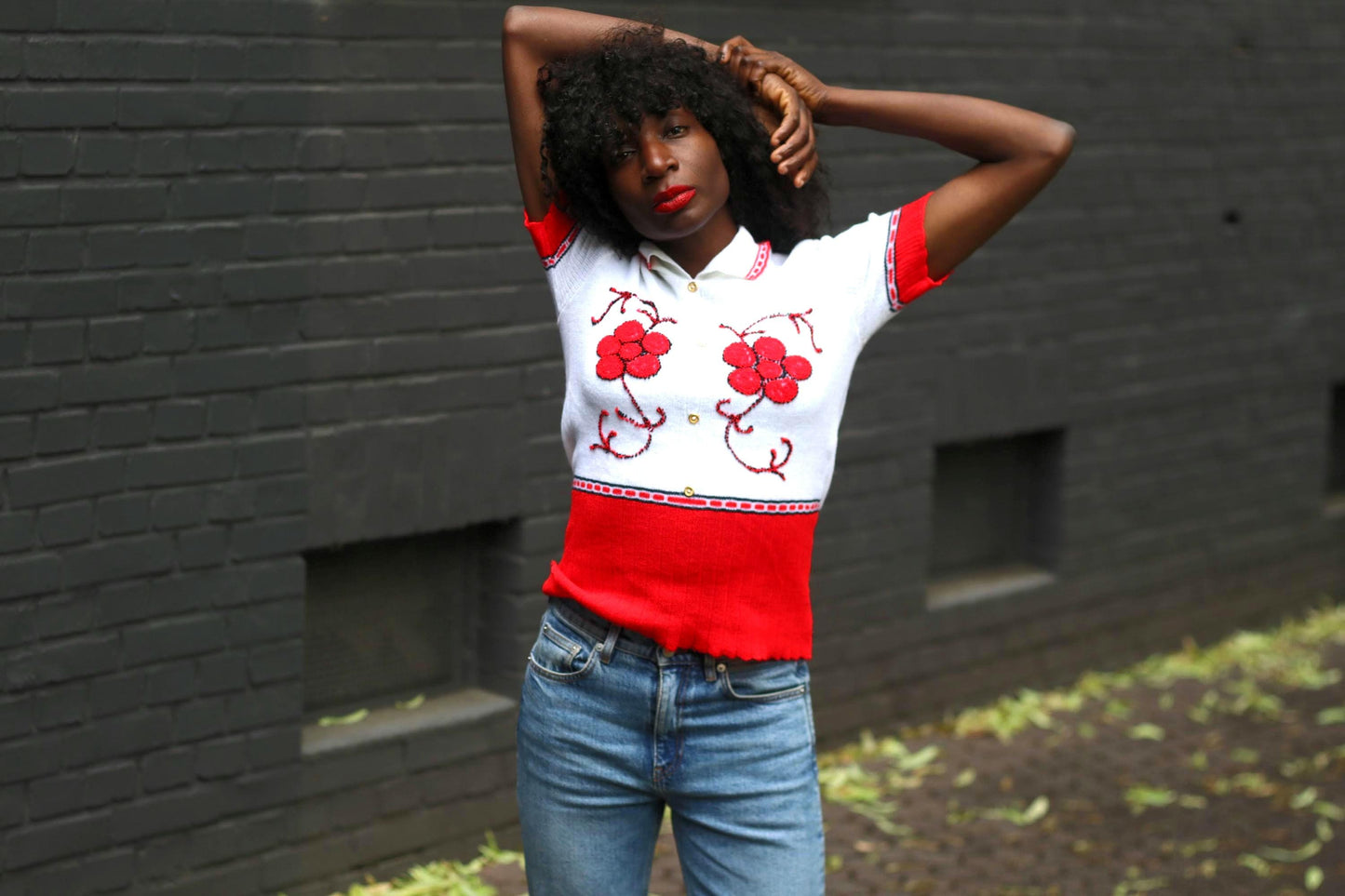 Front view of a woman wearing a vintage red and white knit polo blouse with floral embroidery, hands behind her head, against a dark brick wall.
