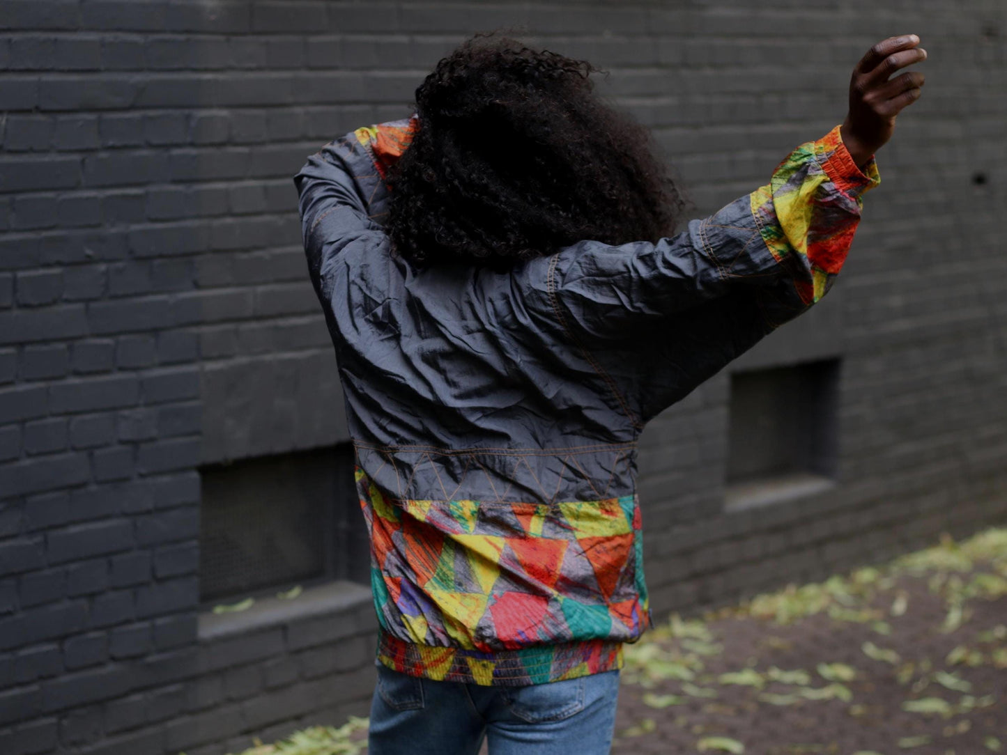 Back view of a female model with curly black hair wearing a vintage 90s windbreaker with colorful hem in front of a gray brick wall. The model's face is away from camera and she has both hands in the air