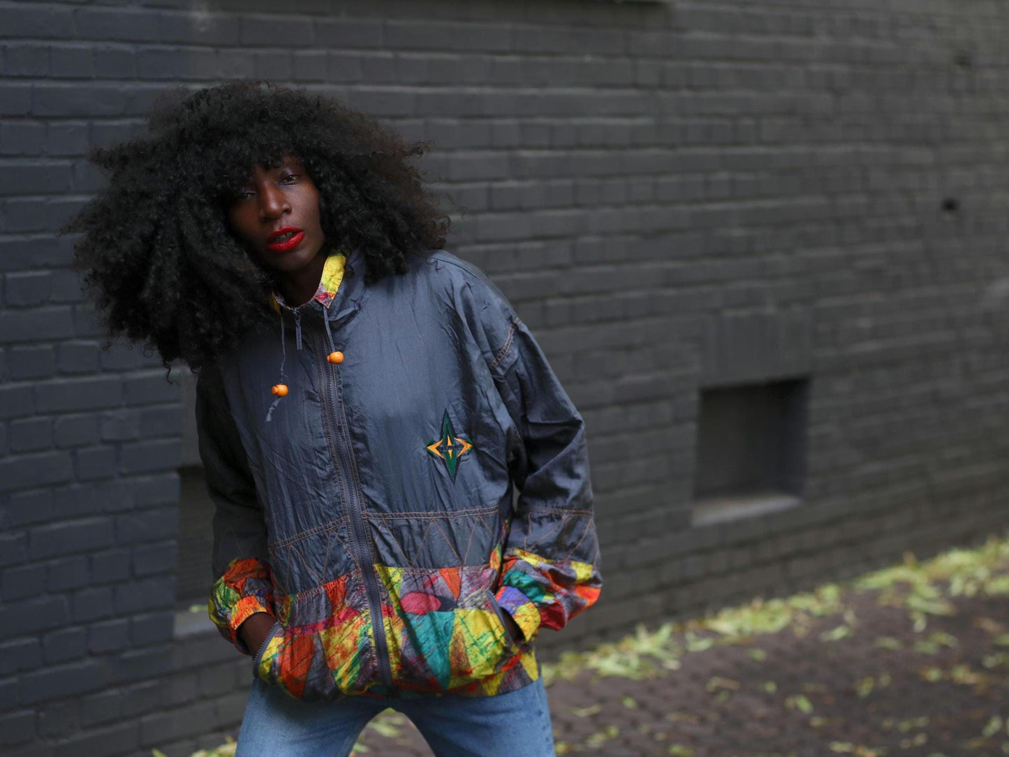 Female model with curly black hair wearing a vintage 90s windbreaker with colorful hem in front of a gray brick wall