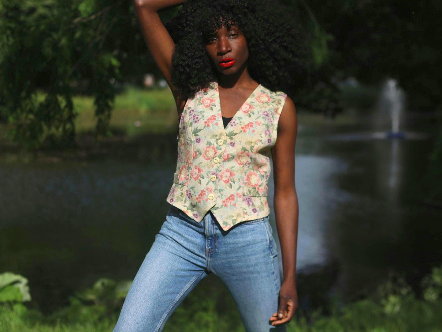 Female model with curly black hair wearing vintage 90s floral vest by a lake. The tapestry handmade vest is styled with blue jeans.