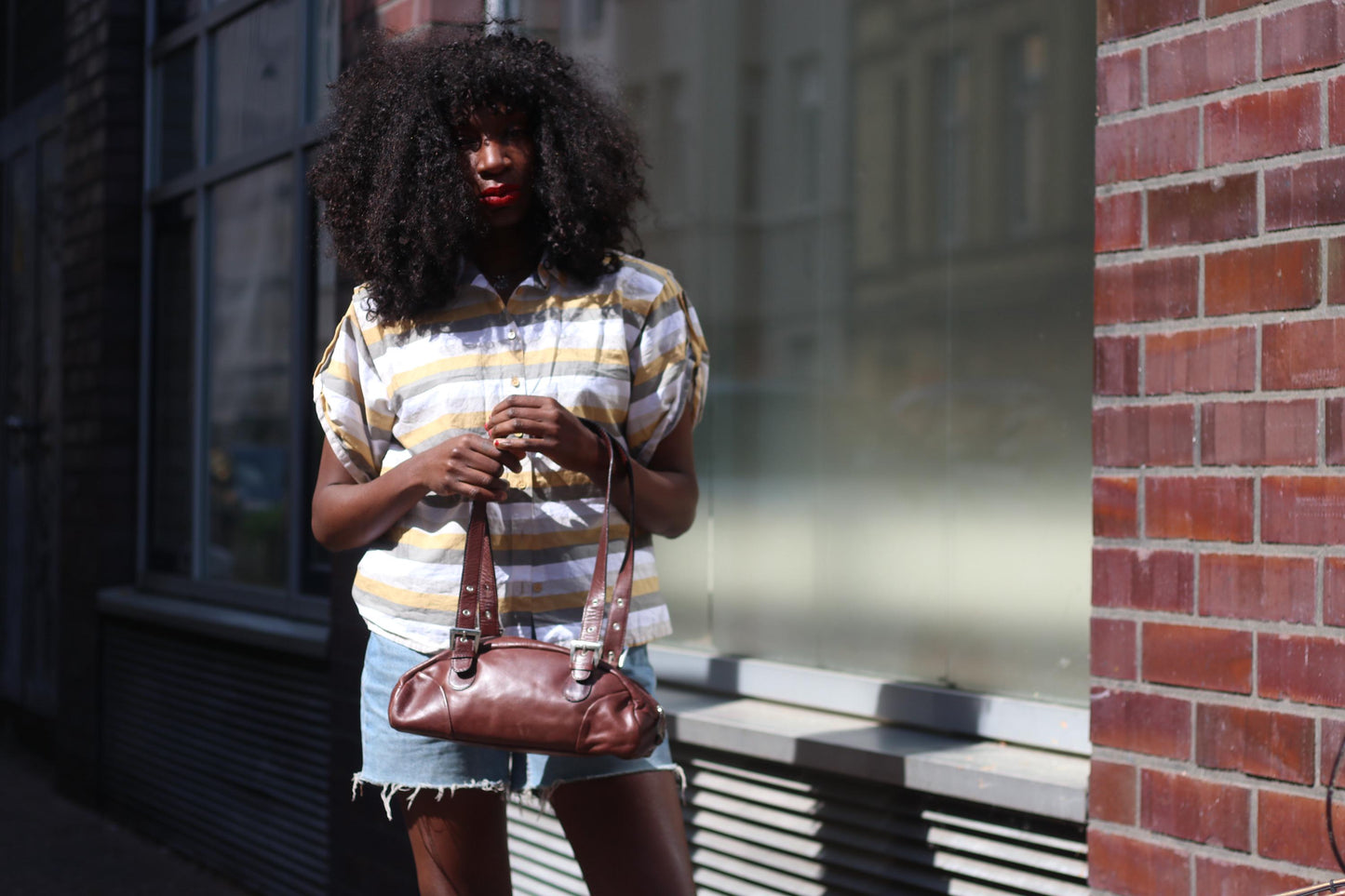 Female model with curly black hair is wearing a striped shirt with blue denim shorts is photographed in an urban setting holding a 90s structured leather bag in rich brown, minimal and elegant with top handles.