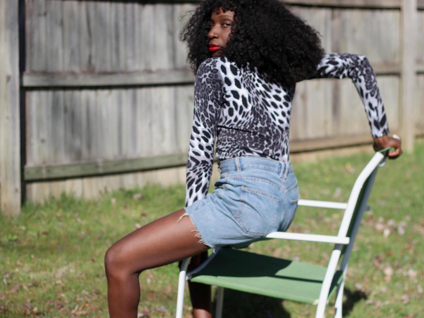 Outdoor portrait of a person wearing a long-sleeved vintage Vertigo Paris shirt with a graphic black and white pattern and denim shorts, sitting on a green lawn chair
