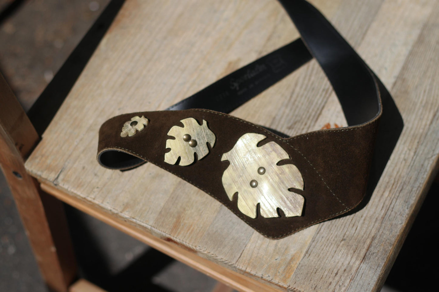 A vintage brown suede leather belt from the 80s with three decorative brass leaf shapes is displayed on a weathered wooden surface.