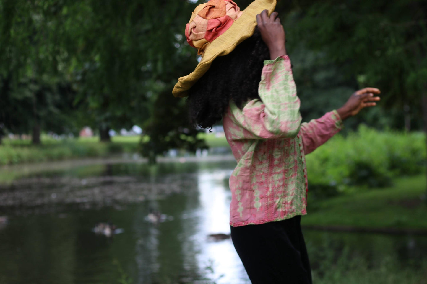Side view of a female model with curly black hair wearing a vintage 90s pink and green tie-dye printed top and large straw hat, looking away from the camera by a pond.