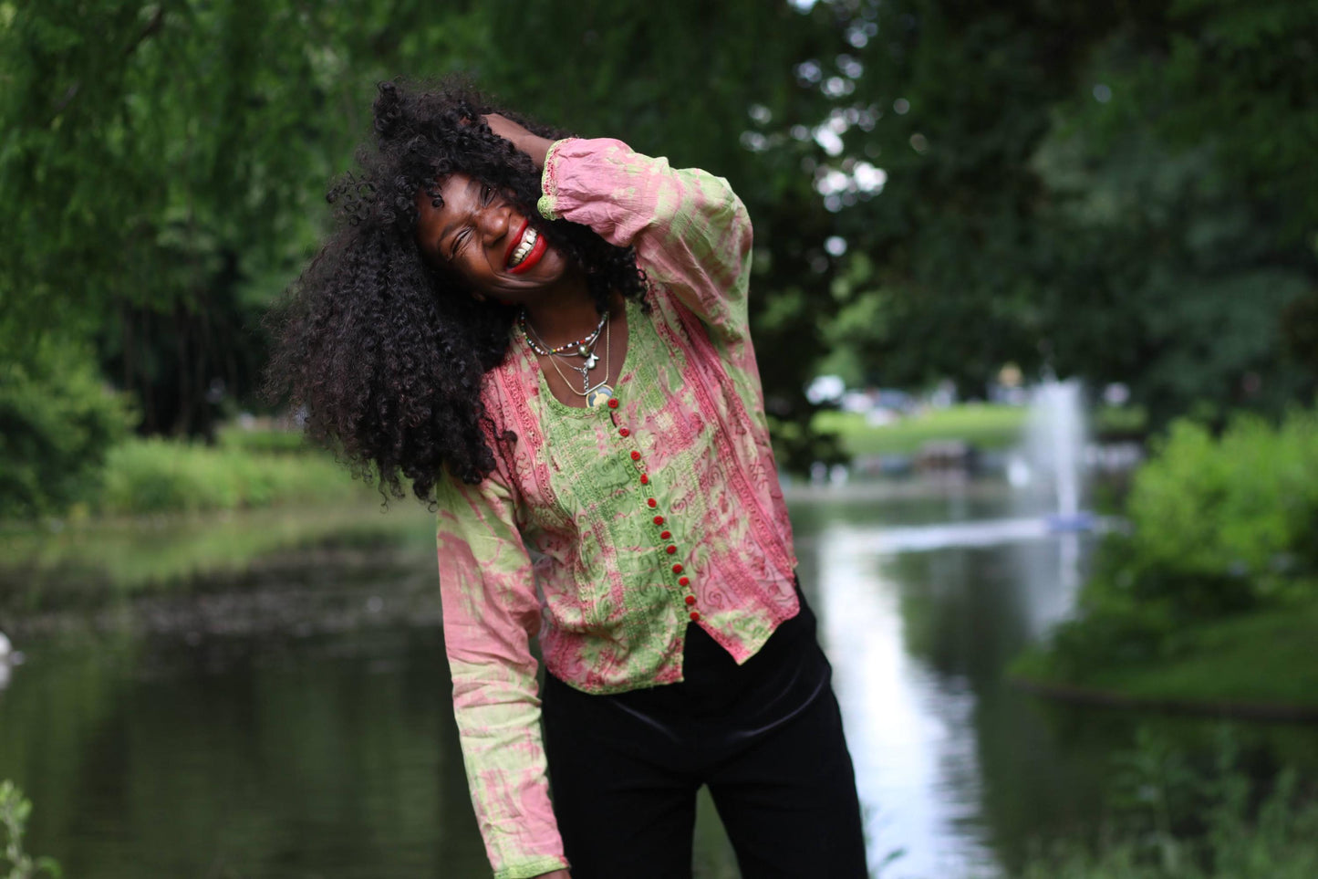 Smiling female model wearing a vintage 90s pink and green tie-dye printed boho chic top and large straw hat, looking into the camera by a pond.