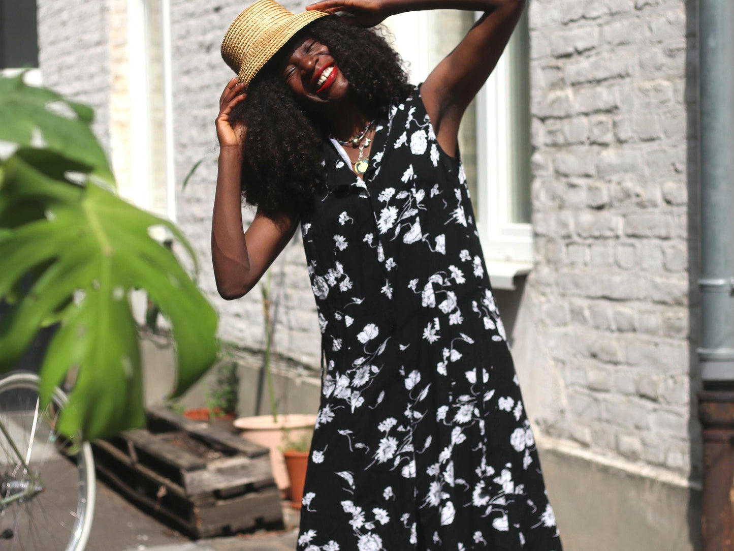 A smiling woman wearing a black sleeveless dress with white floral patterns and a wide-brimmed straw hat poses outdoors, looking directly into the camera