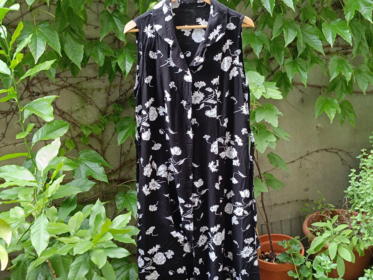full-length shot of a retro vintage black sleeveless dress with white floral patterns, with green foliage partially obscuring the dress on the left side. The fabric drapes loosely.