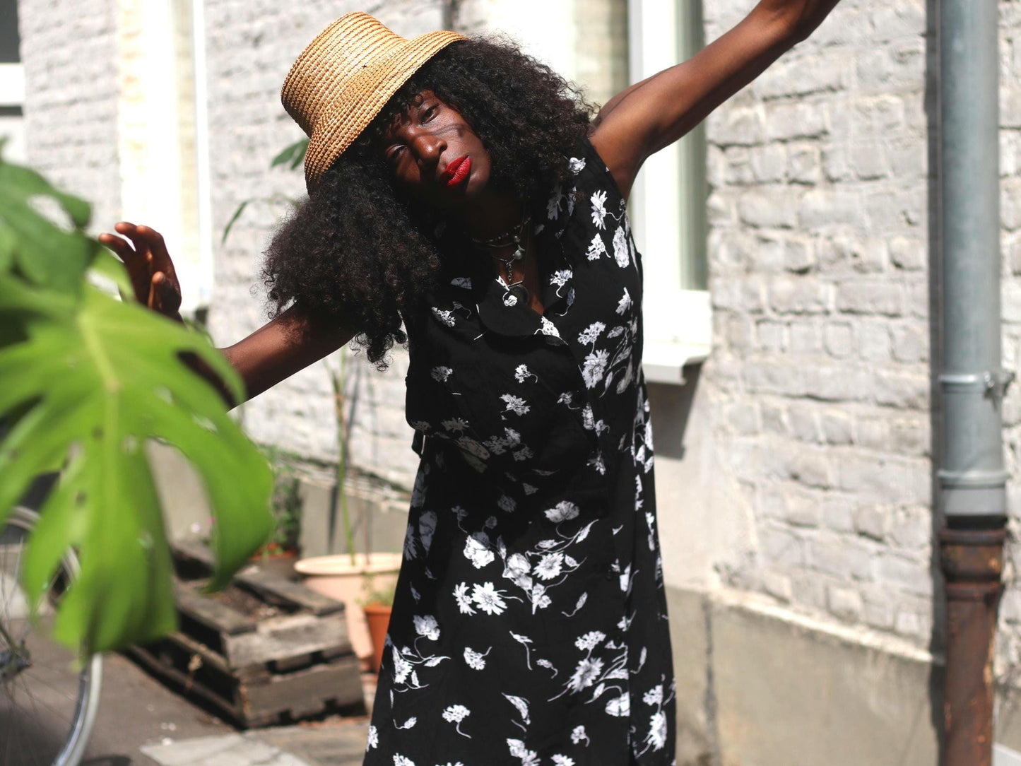 A woman with dark curly hair wearing a vintage black sleeveless dress with white floral patterns stands outdoors, holding a large green leaf. She is looking away from the camera, with the dress flowing around her.