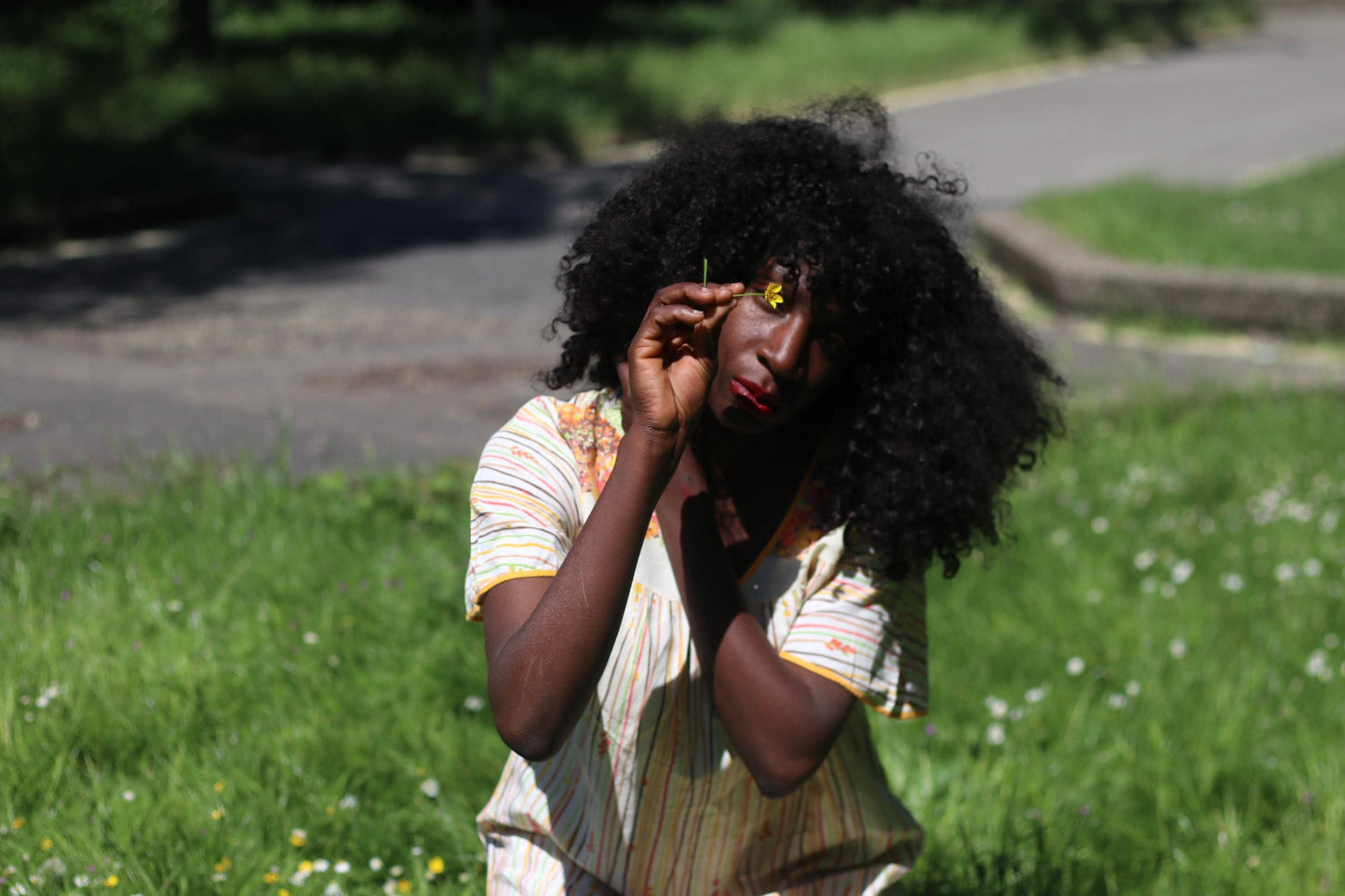 A female model with curly hair sits in a field of grass in a boho retro style 70s dress and holds a single small yellow flower close to her eyes