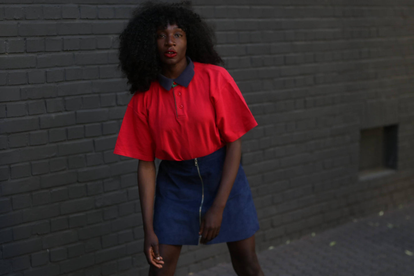A female model with dark, voluminous curly hair wearing a bright red collared shirt and a dark blue suede skirt with a front zipper, against a textured gray brick wall.