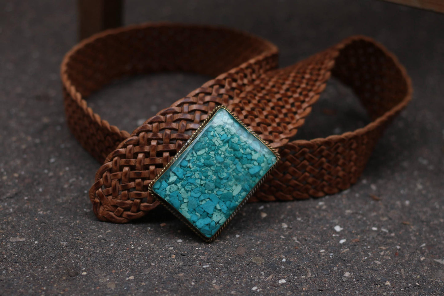 Coiled vintage braided brown leather belt with the turquoise buckle in the center is photographed on a grey asphalt surface