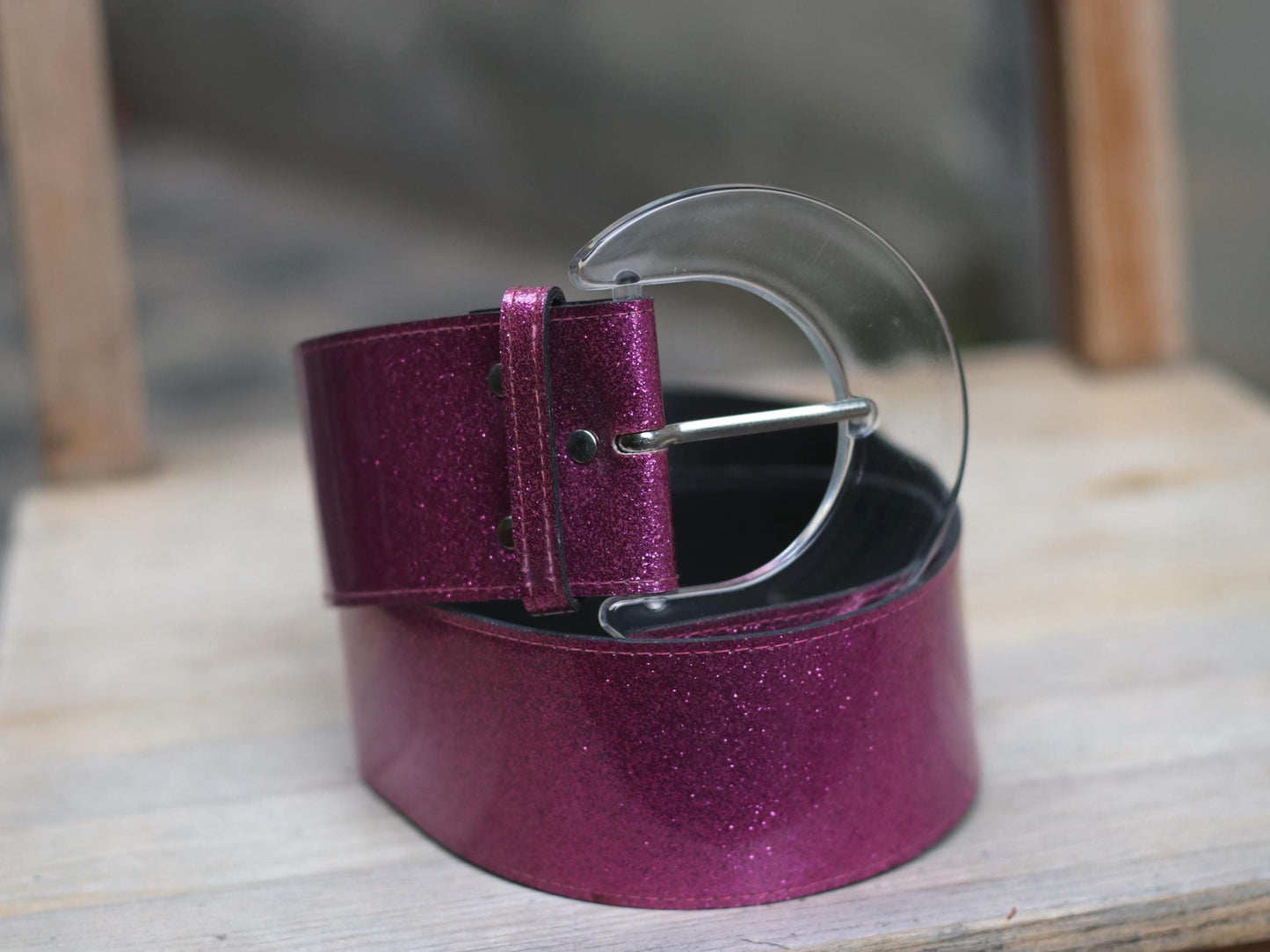 A close-up of a Y2K-style belt featuring a vibrant magenta glitter strap layered over a wide black leather strap, secured by a clear half-moon buckle, on a wooden surface.