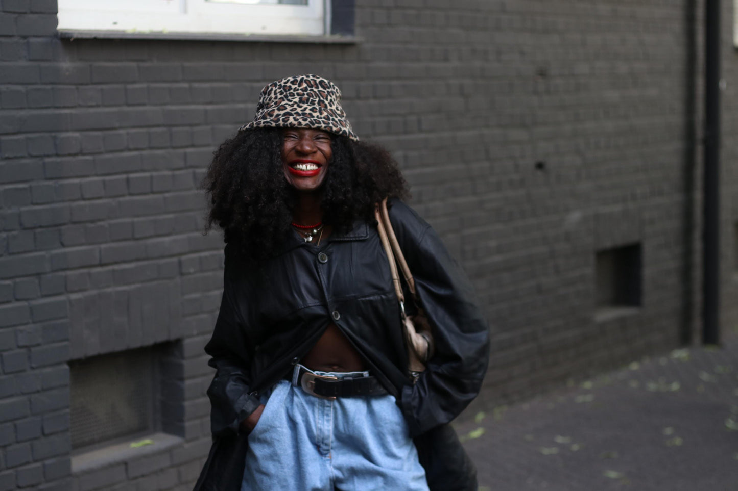 A Vintage black leather jacket styled on a smiling female model wearing a patterned hat and tall animal print boots, photographed against a grey wall.