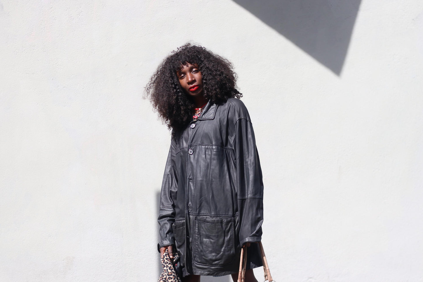 A female with curly hair photographed against a sunlight white wall outdoors wearing an oversized vintage black 90s leather jacket and denim shorts