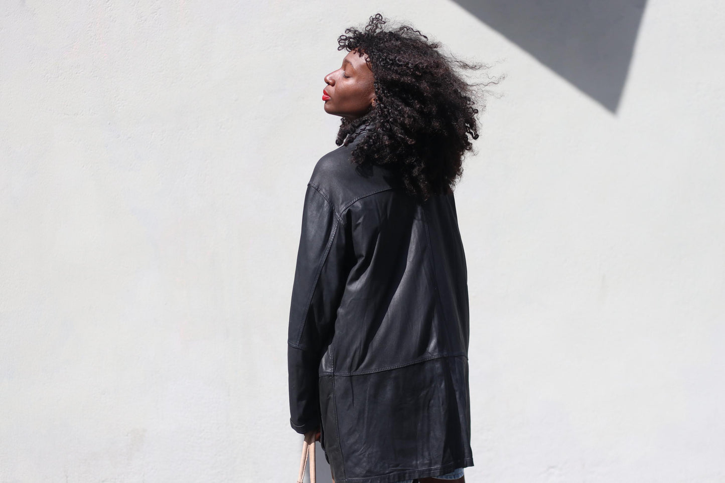 A side view of a female model with curly hair wearing a black 90s leather trench jacket, denim shorts standing outdoors.