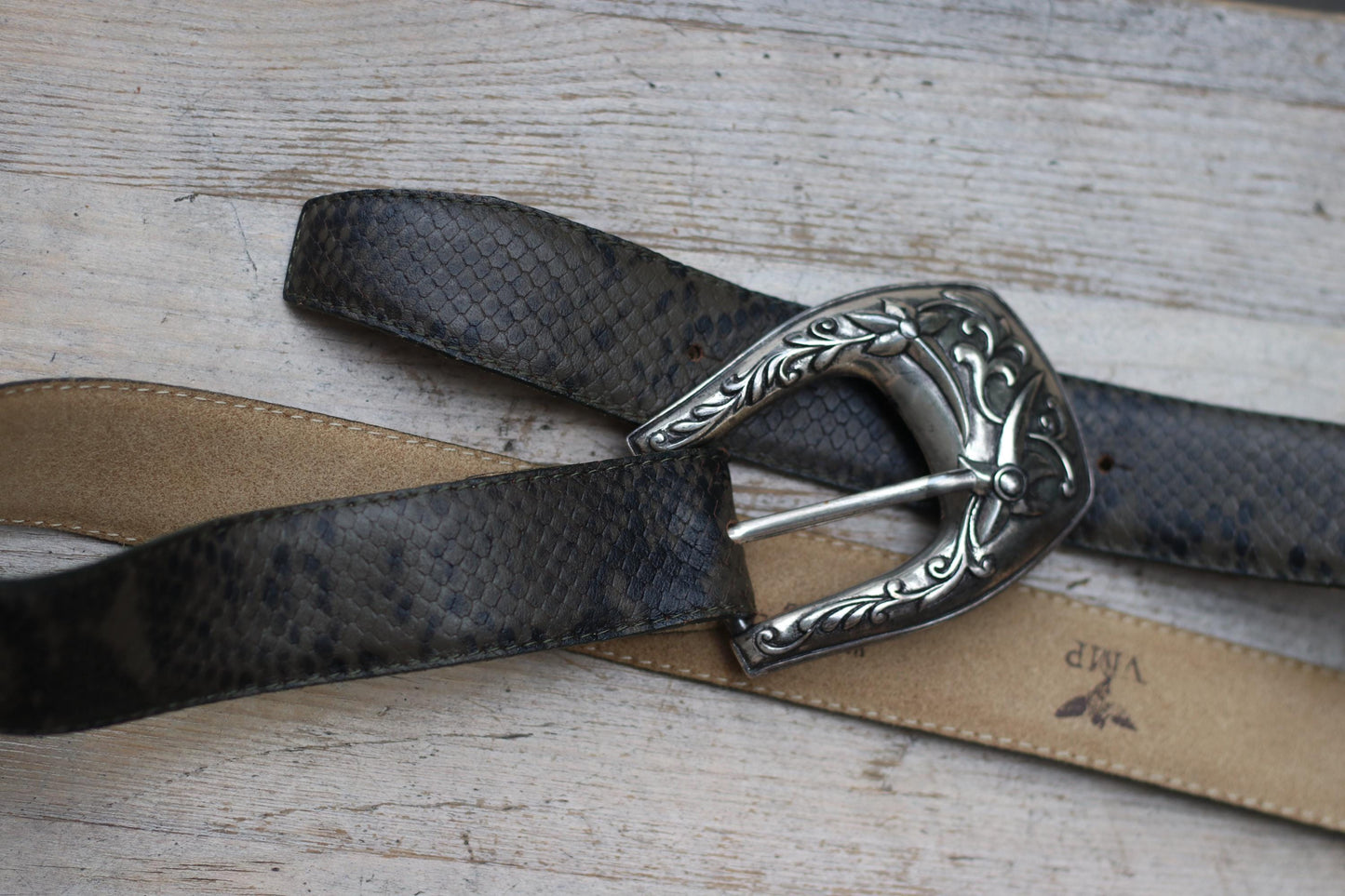 Overhead shot of a dark snakeskin-embossed vintage western belt with an ornate silver buckle, lying on a light wooden surface