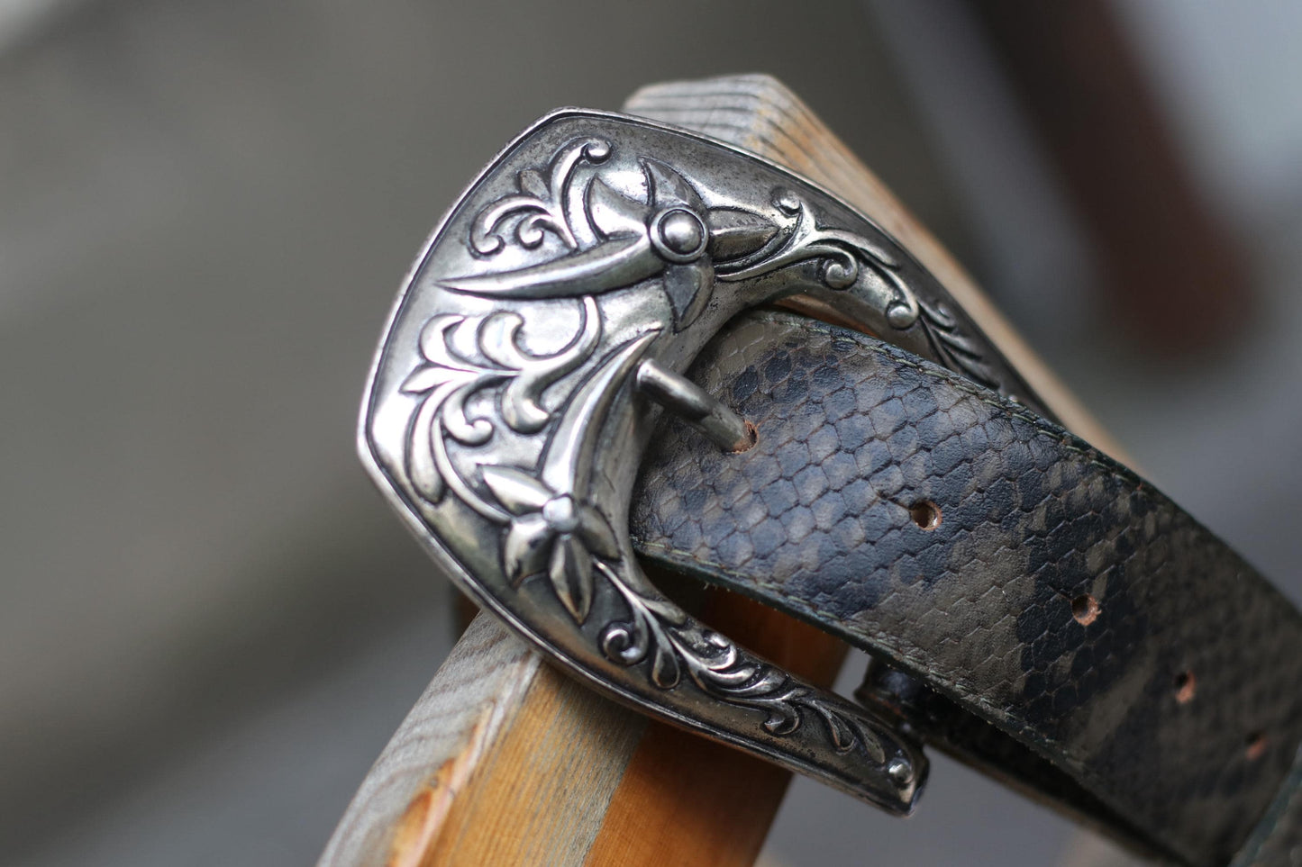 Close-up of a vintage ornate silver western belt buckle with intricate floral and scroll engravings, attached to a dark brown snakeskin-embossed leather belt