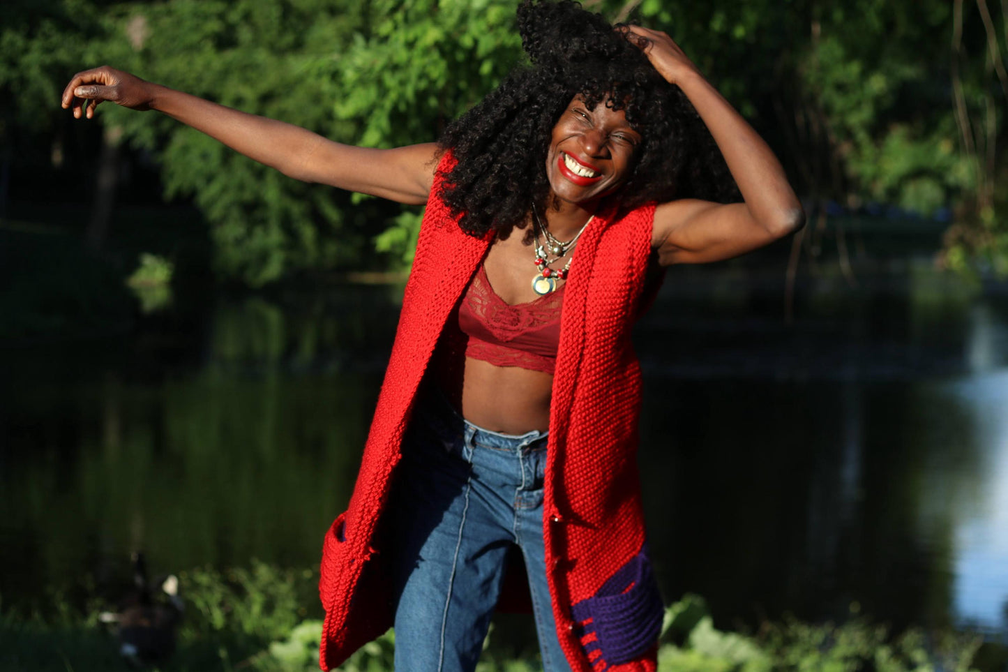 Smiling female model with curly hair is photographed outdoors wearing a red vintage knitted vest with purple pockets over a red cropped top with lace trimmings.