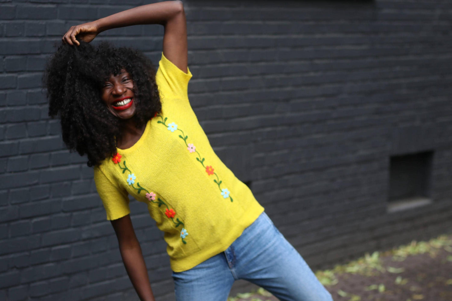 Cheerful 70s-inspired outfit featuring a female model with curly black hair in a retro sunny vintage embroidered tee and casual blue jeans in an outdoor setting.