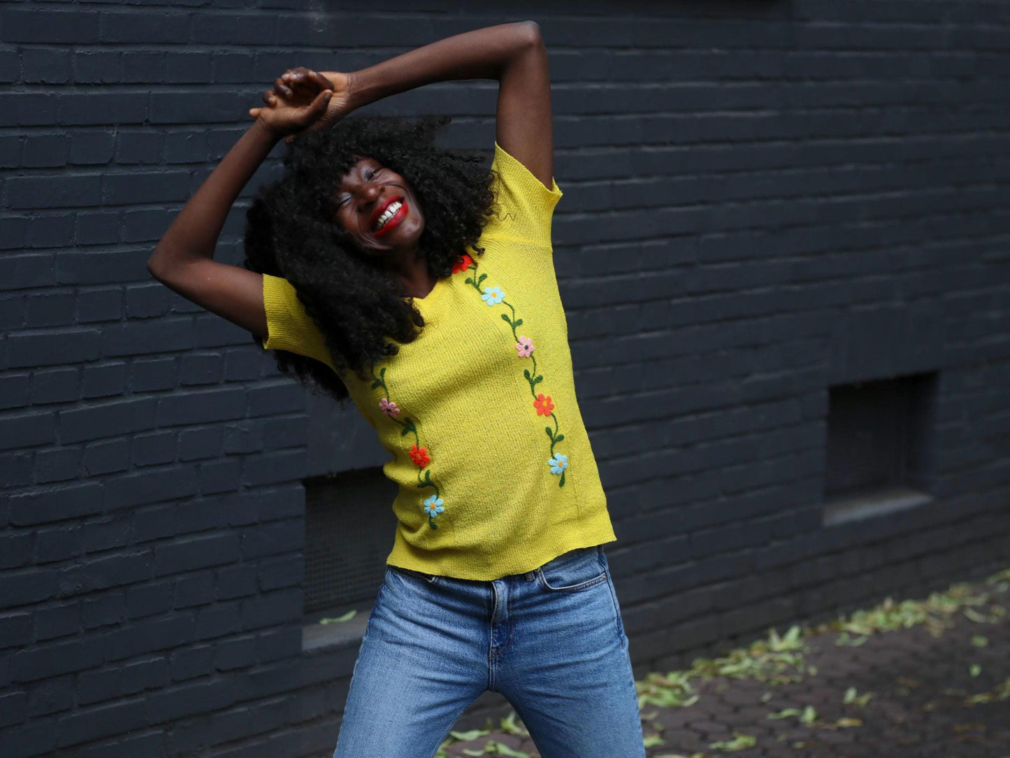 Smiling woman wearing a vintage yellow knit top with floral embroidery, posing joyfully against a dark brick wall.