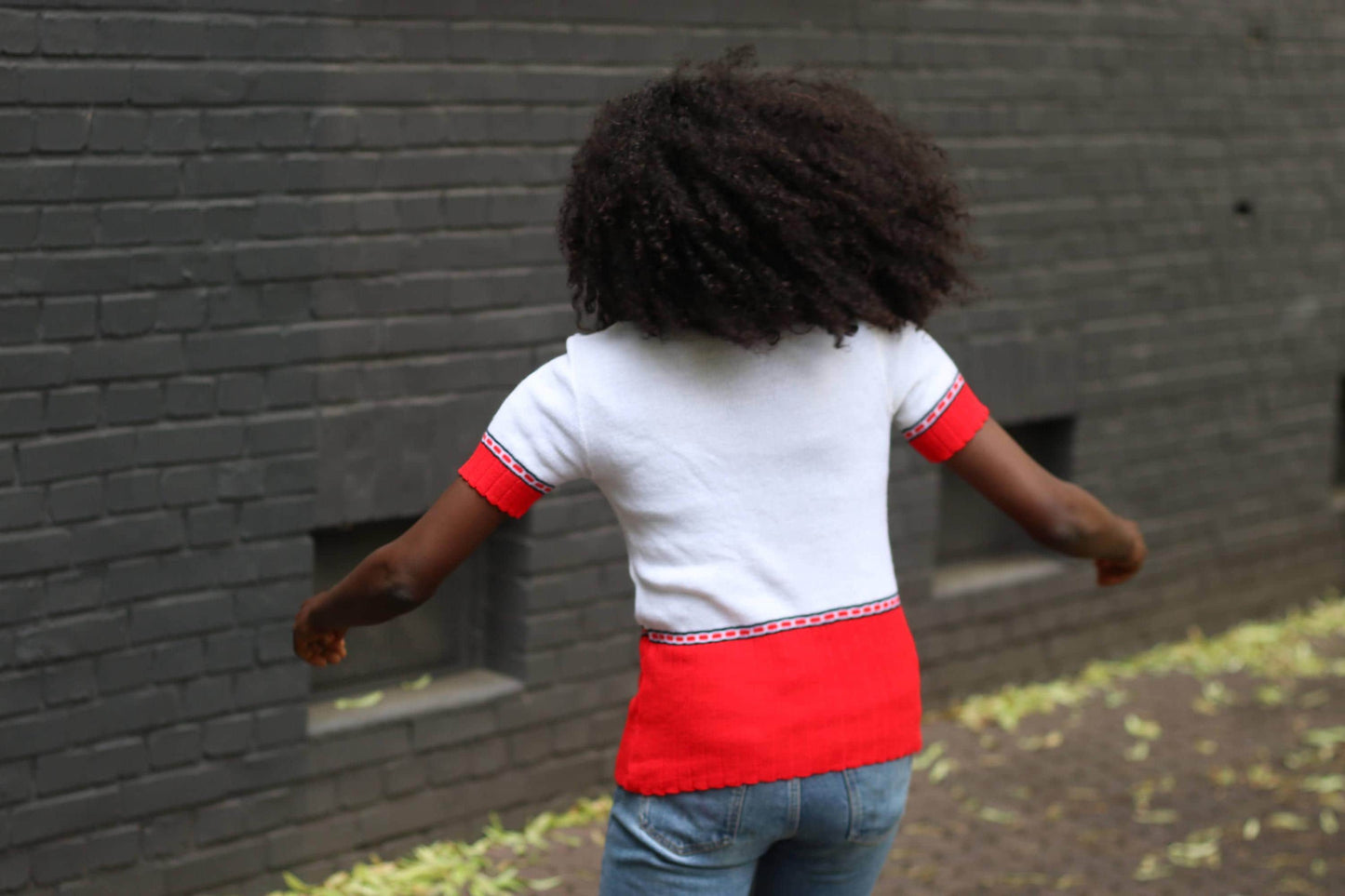 Back view of a female model person in a white and red knit vintage polo shirt with short sleeves, standing in front of a dark brick wall