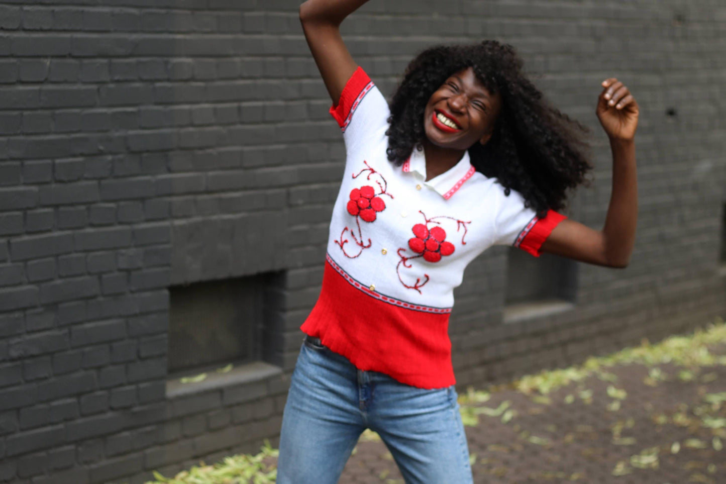 Full shot of a female model in a vintage red and white knit polo shirt with embroidered flowers, paired with blue jeans, posing joyfully outdoors.