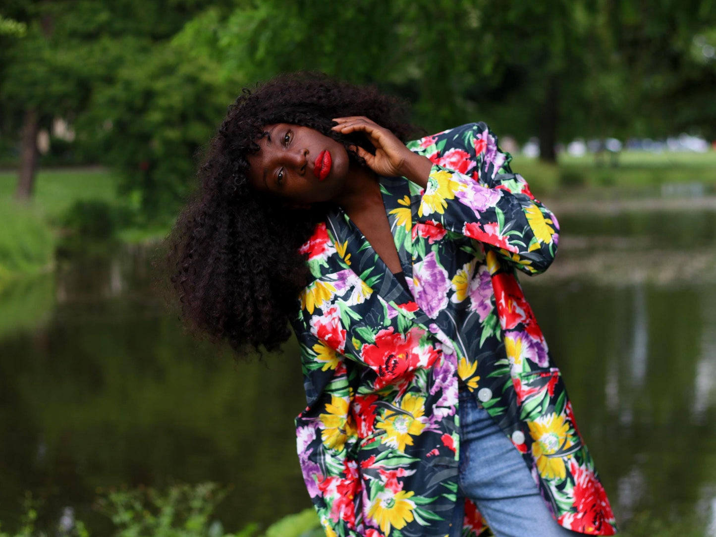 Female model with curly black hair wearing a vintage 90s floral print blazer with blue denim, photographed outdoors infron of a lake. The oversized floral jacket is perfect for retro tropical style with a modern edge.