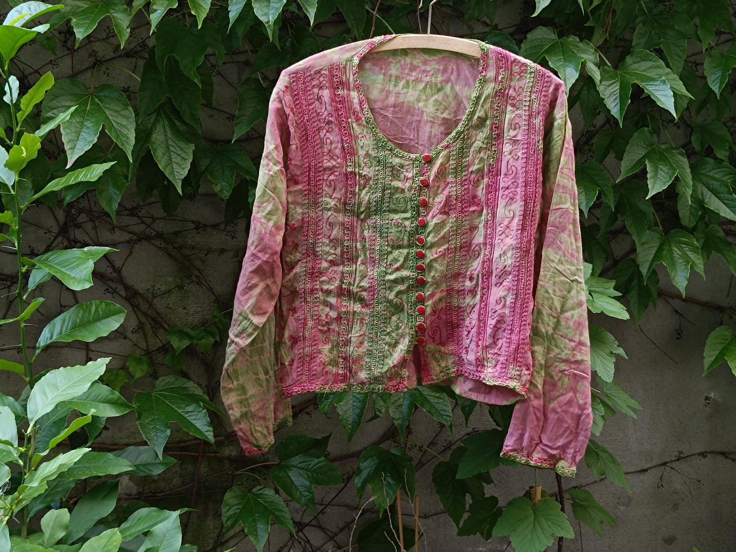 Vintage tie-dye boho blouse with pink and green patterned top photographed hanging outdoors on a clothing line with leaves in the background. The still life image showcases the print and texture of the retro long-sleeved blouse.