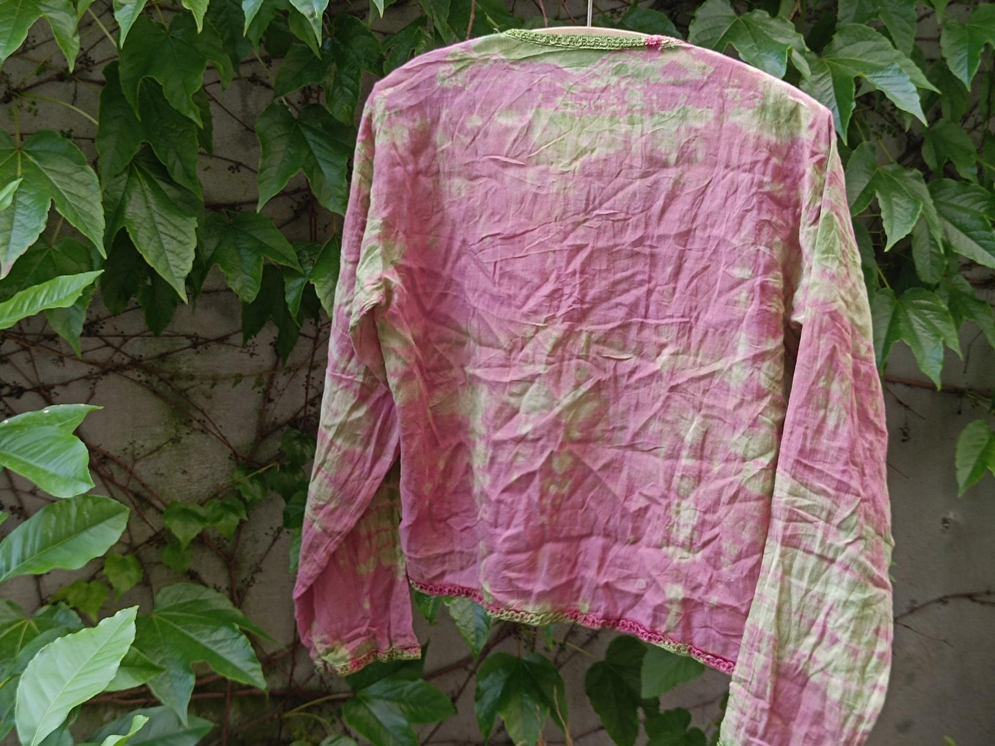 Back view of a vintage boho chic tie-dye blouse with pink and green colors photographed hanging outdoors on a clothing line with leaves in the background. The still life image showcases the print and texture of the retro long-sleeved blouse.